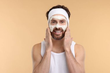 Saç bandı takmış bir adam bej arka planda yüzünü yıkıyor.