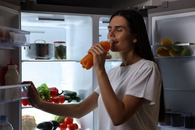 Geceleri modern buzdolabının yanında meyve suyu içen genç bir kadın.