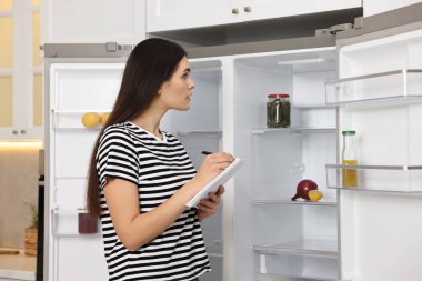 Genç bir kadın mutfaktaki boş buzdolabının yanına notlar yazıyor.