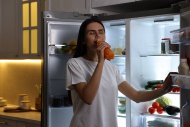 Geceleri modern buzdolabının yanında meyve suyu içen genç bir kadın.