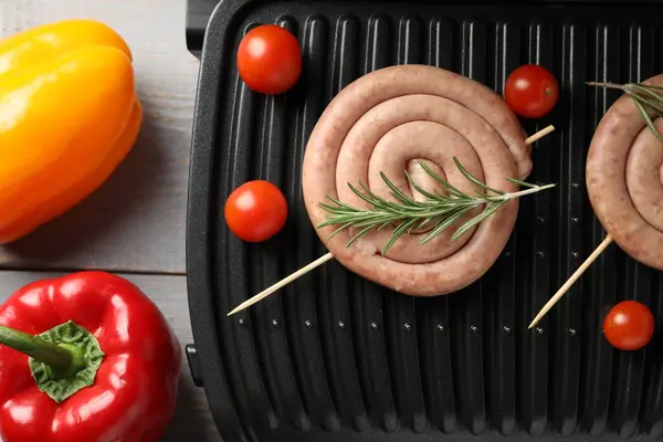 Electric grill with homemade sausages, rosemary and vegetables on rustic wooden table, flat lay