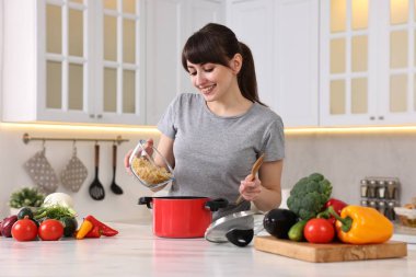 Mutfaktaki beyaz mermer masada çömleğe çiğ makarna ekleyen genç ve mutlu ev hanımı. Yemek pişirme süreci