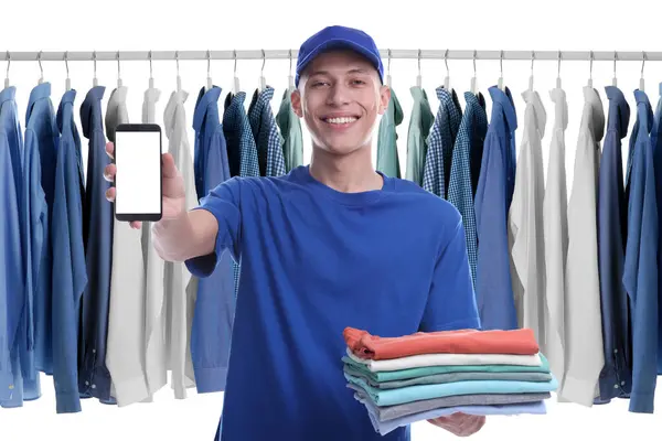 Dry-cleaning delivery. Happy courier holding folded clothes and smartphone near wardrobe rack on white background