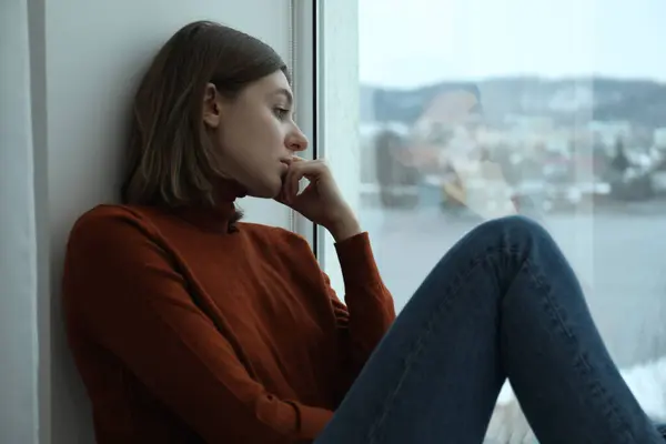 stock image Sad young woman sitting on windowsill near window at home