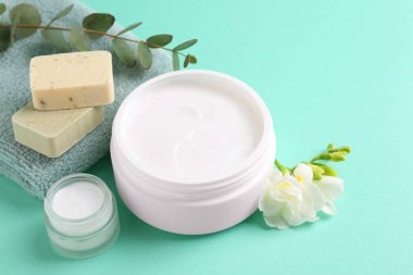 Açık kavanozlarda nemlendirici krem, sabun barları ve turkuaz arka planda frezya çiçeği. Vücut bakım ürünleri