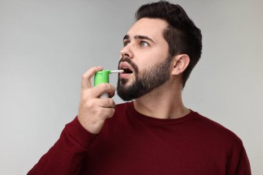 Gri arka planda boğaz spreyi kullanan genç adam.
