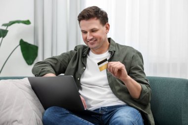 Kredi kartı olan mutlu bir adam evdeki kanepede online alışveriş için dizüstü bilgisayar kullanıyor.