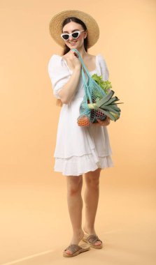 Bej arka planda taze sebzelerle dolu bir sicim çantası olan kadın.