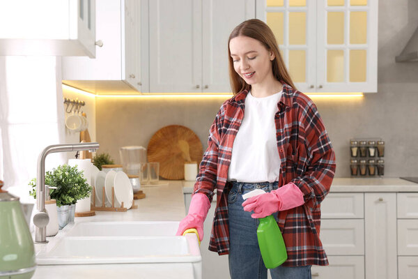 Woman with spray bottle and microfiber cloth cleaning sink in kitchen