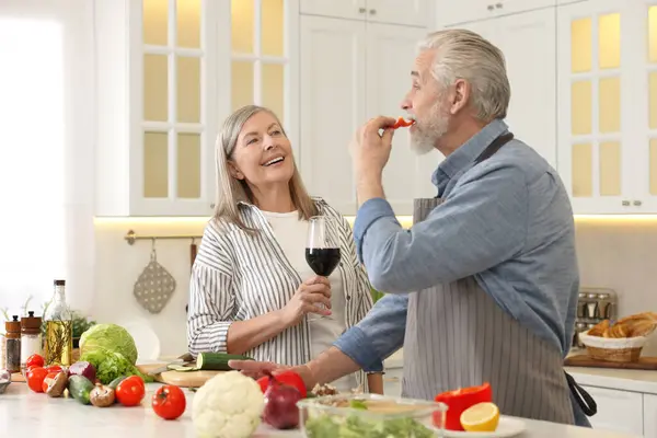 Happy senior couple cooking together in kitchen. Woman with glass of wine near her husband