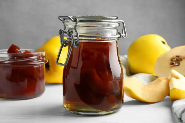 Tasty homemade quince jam in jars and fruits on white wooden table, closeup