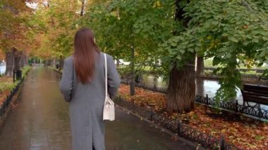 Gri ceketli çekici bir kadın yağmurdan sonra sonbahar parkında yürüyor. Kamera onun peşinden gidiyor.