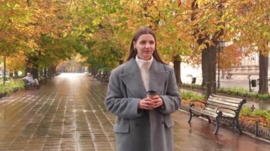 Gri ceketli çekici bir kadın yağmurdan sonra sonbahar parkında yürürken sıcak bir şeyler içer. Kamera onunla hareket ediyor.