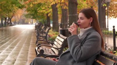 Gri ceketli çekici bir kadın sonbahar parkında bankta otururken sıcak içkisinin tadını çıkarıyor.