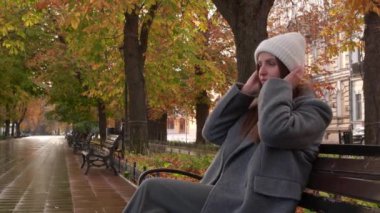 Gri ceketli ve ılık beyaz şapkalı çekici bir kadın sonbahar parkında bankta oturuyor.