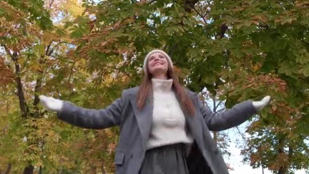 スタイリッシュな灰色のコートで魅力的な女性 暖かい白い帽子と秋の公園で楽しんでいるミトン 低角度ビュー — ストック動画