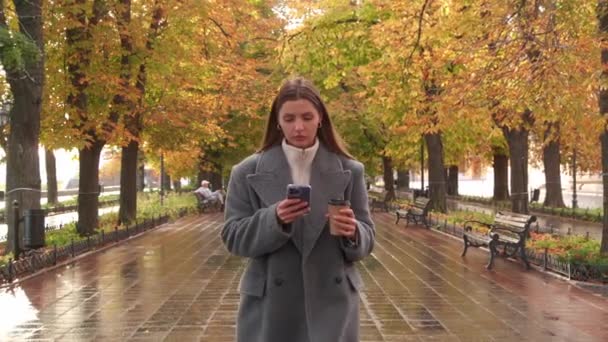 迷人的女人穿着时髦的灰色外套 带着外卖热饮 雨后在秋天公园散步时使用手机 相机和她一起移动 — 图库视频影像