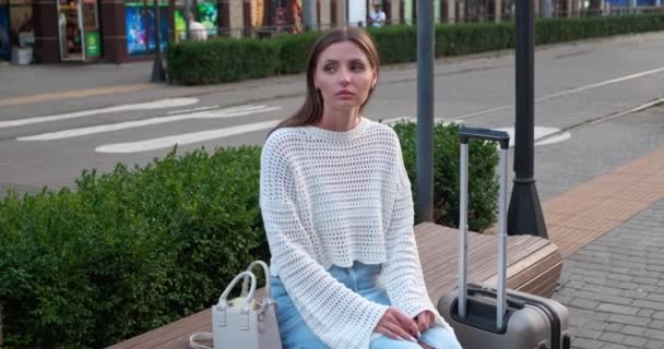 美しい若い女性はベンチに座って 屋外でタクシーを待っている間電話を使用しています — ストック動画