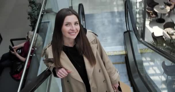 在商场乘自动扶梯时 提着购物袋看着相机 笑着的年轻女子 — 图库视频影像