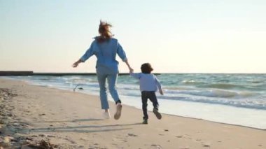 Anne oğluyla el ele tutuşup gün batımında deniz kenarında koşuyor. Kamera izleme