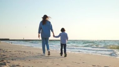 Anne oğluyla el ele tutuşup gün batımında deniz kenarında yürüyor. Kamera izleme