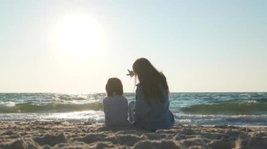 Anne ve oğlu gün batımında, deniz kenarında sarılıyorlar.