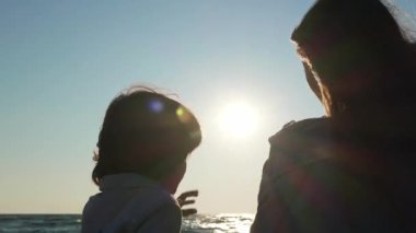 Anne ve oğlu gün batımında, deniz kenarında elleri ile kalp yapıyor. Kamera hareket ediyor, parmaklarının arasından güneşi gösteriyor.
