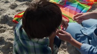 Anne ve oğlu, kumların üzerinde uçurtma dizisi kuruyorlar. Kamera onların etrafında hareket ediyor.