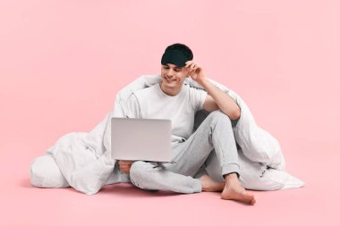 Pembe arka planda uyku maskesi, battaniyesi ve laptopu olan mutlu bir adam.