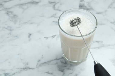 Mini mixer (milk frother) and tasty cappuccino in glass on white marble table, closeup. Space for text clipart