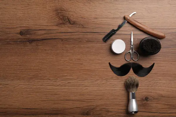 Artificial moustache and barber tools on wooden table, flat lay. Space for text