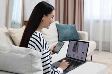 Akıllı telefonu ve dizüstü bilgisayarı olan güzel genç bir kadın evde online alışveriş yapıyor.