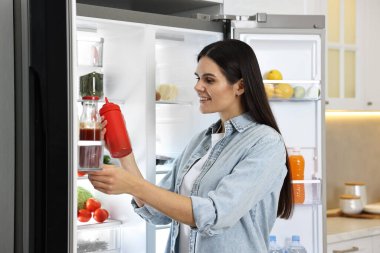 Genç bir kadın mutfaktaki buzdolabından ketçap alıyor.