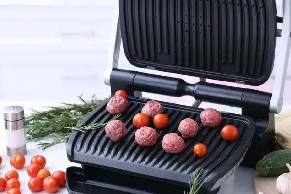 Electric grill with meatballs, tomatoes and rosemary on white table