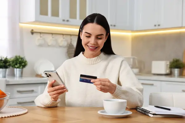 stock image Happy young woman with smartphone and credit card shopping online at wooden table in kitchen