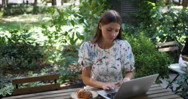 Açık kafedeki ahşap masada dizüstü bilgisayar kullanan düşünceli bir kadın.