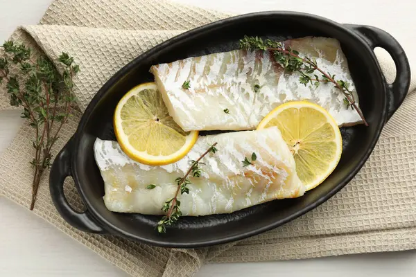 Fresh raw cod fillets with thyme and lemon in baking dish on white wooden table, top view