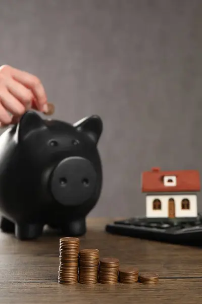 Woman putting coin into piggy bank, closeup. House model, calculator and money on wooden table, selective focus