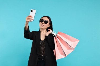 Alışveriş torbalarıyla gülümseyen kadın açık mavi arka planda selfie çekiyor.