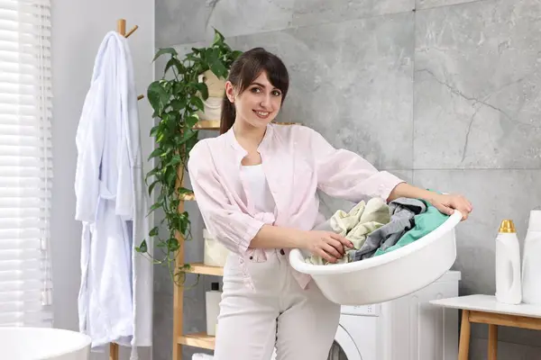 stock image Happy young housewife holding basin with laundry in bathroom