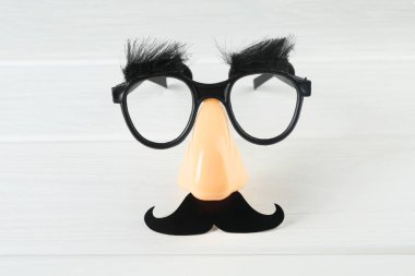 Takma bıyıklı komik bir maske, beyaz ahşap arka planda burun ve gözlük.