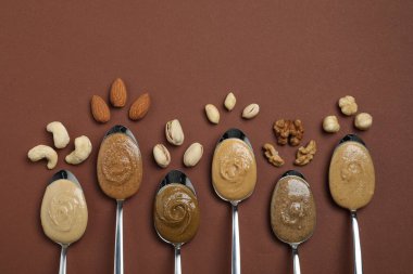 Tasty nut butters in spoons and raw nuts on brown table, flat lay. Space for text clipart