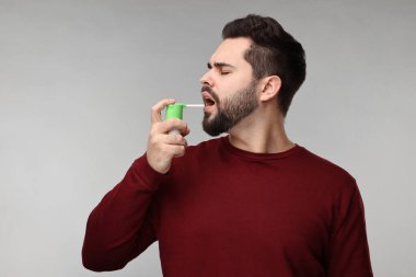 Gri arka planda boğaz spreyi kullanan genç adam.