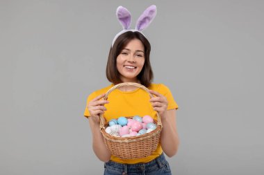 Paskalya kutlaması. Tavşan kulaklı mutlu kadın ve gri arka planda boyanmış yumurta dolu hasır sepet.