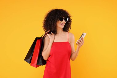 Alışveriş torbaları ve sarı arka planda modern akıllı telefonları olan mutlu genç bir kadın.