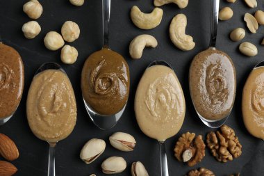 Tasty nut butters in spoons and raw nuts on dark gray table, flat lay clipart