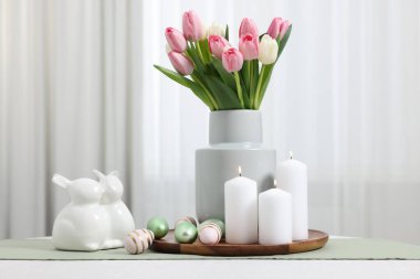 Paskalya süsleri. Bir buket lale, boyalı yumurta, yanan mumlar ve beyaz masa üzerinde tavşan figürleri.