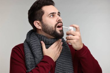Gri arka planda boğaz spreyi kullanan atkılı genç bir adam.