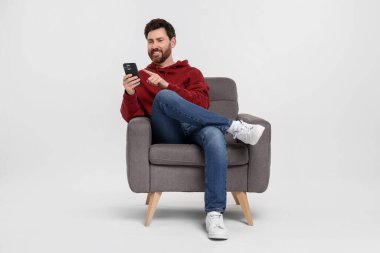 Akıllı telefonlu mutlu bir adam beyaz arka planda koltukta oturuyor.