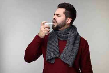 Gri arka planda boğaz spreyi kullanan atkılı genç bir adam.